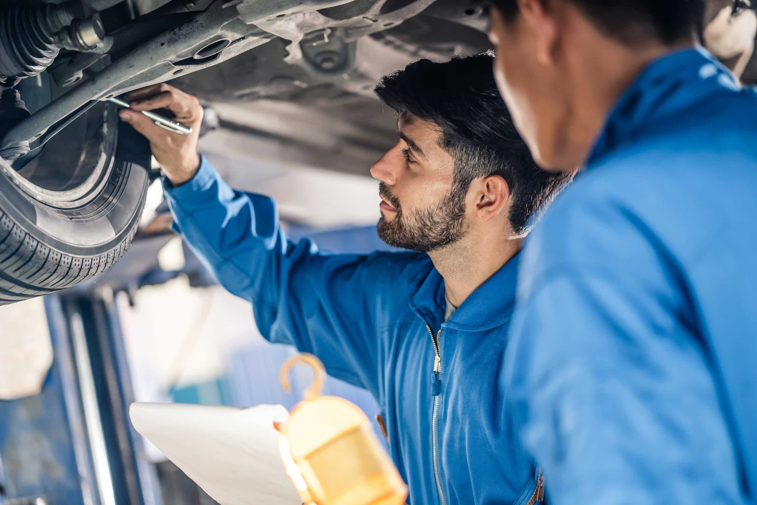 Vehicle service maintenance handsome mens checking under car condition in garage. Automotive mechanic pointing flash light on wheel following maintenance checklist document. Car repair service concept